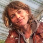 Eine Frau mit braunem, gewelltem Haar und einem warmen Lächeln trägt eine braune Lederjacke und einen grauen Hoodie. Sie ist im Freien, mit gestreiften Markisen im Hintergrund.