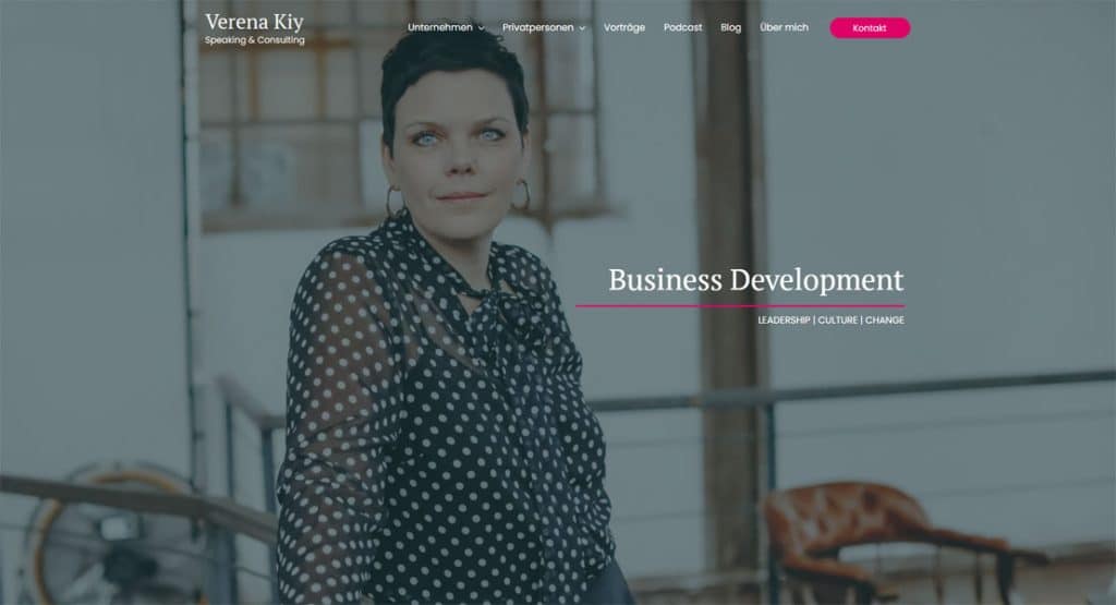 Verena Kjy, eine Frau mit kurzen Haaren und einem gepunkteten Oberteil, blickt nachdenklich in die Kamera. Im Hintergrund ist ein unscharfes Gebäude zu sehen. Der Text "Business Development" ist prominent platziert.